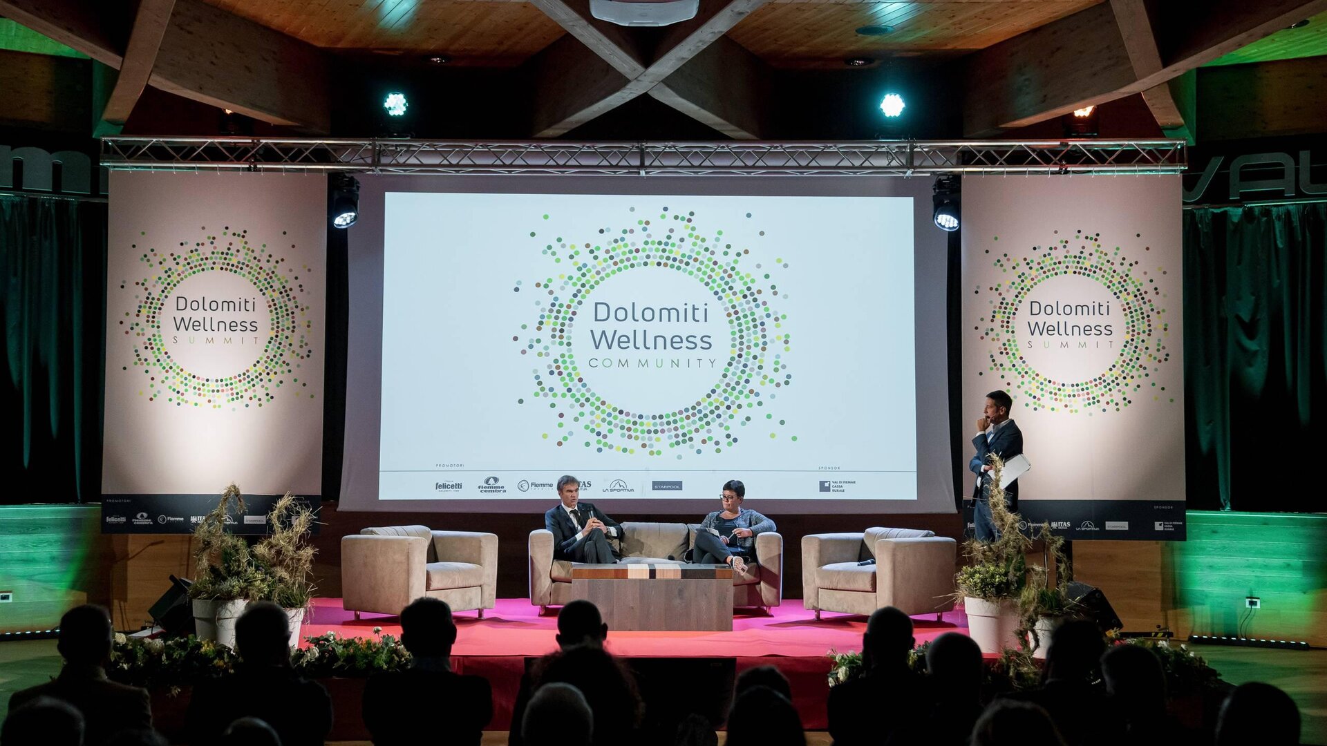 Un evento di wellness nelle Dolomiti, con relatori su un palco attrezzato. Il pubblico assiste mentre viene presentato il tema "Dolomiti Wellness Community".