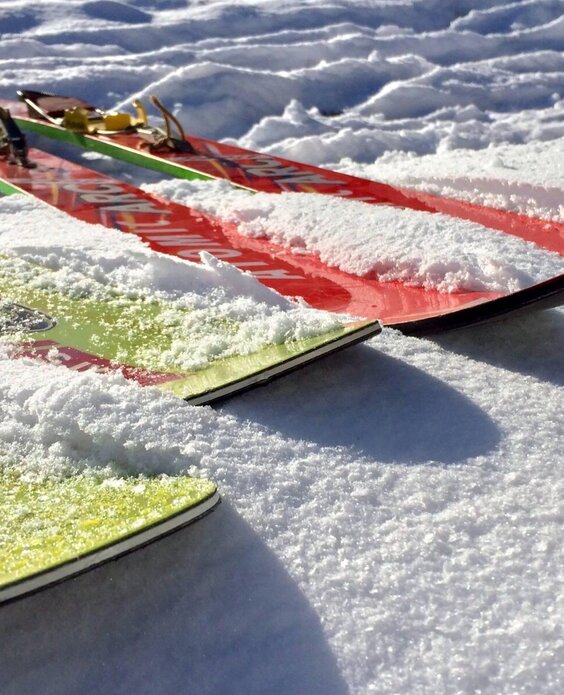 Due sci colorati pendono da un seggiovia su una pista innevata. In lontananza si vedono il cielo azzurro e alcune piante.