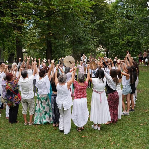 Un gruppo di persone si raduna in un parco, sollevando le mani verso il cielo. Indossano abiti bianchi e colorati, creando un'atmosfera di festa e comunità.