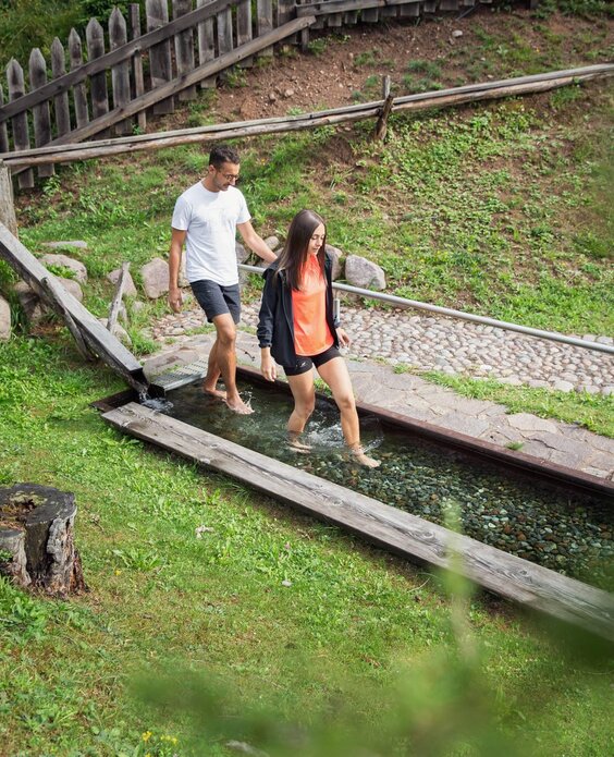 A couple walks barefoot on a wooden path in a natural setting. Around them, there are trees and a wooden fence.