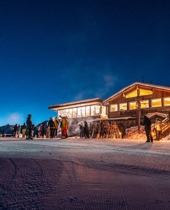 Una baita illuminata di sera circondata da neve e sciatori. Il cielo è blu scuro e limpido.