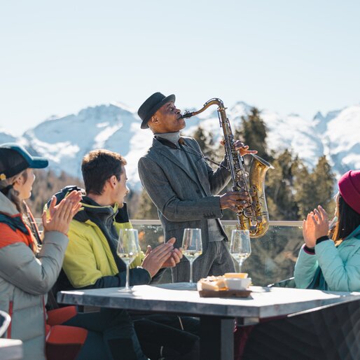 James Thompson suona il sax mentre dei ragazzi fanno aperitivo e lo applaudono | © APT Fiemme e Cembra - Gaia Panozzo