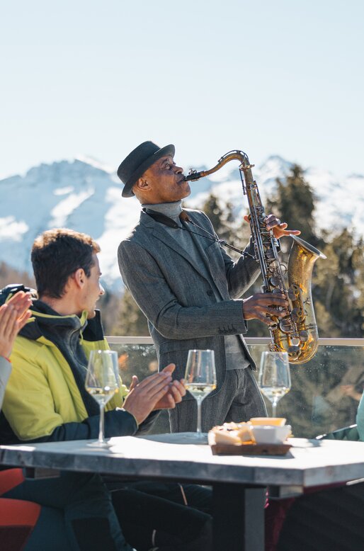 James Thompson suona il sax mentre dei ragazzi fanno aperitivo e lo applaudono | © APT Fiemme e Cembra - Gaia Panozzo