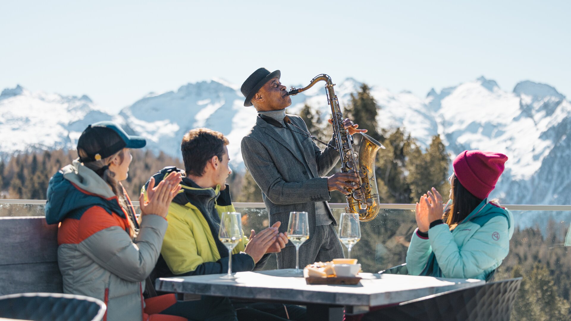 James Thompson suona il sax mentre dei ragazzi fanno aperitivo e lo applaudono | © APT Fiemme e Cembra - Gaia Panozzo