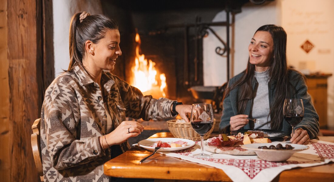 Due amiche condividono un tagliere di salumi e delle castagne, accompagnando il tutto con un bicchiere di vino rosso | © APT Fiemme e Cembra - foto di Gaia Panozzo